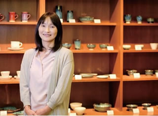 陶芸作品の並ぶ棚の前に立っている山岡みどりさんの写真