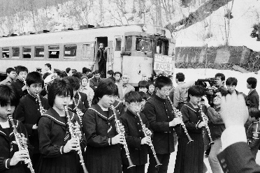 制服を着た栗沢中学校の吹奏楽部の生徒たちが楽器を手に演奏し、背後に万字線の鉄道車両が写る、さよなら万字線の白黒写真