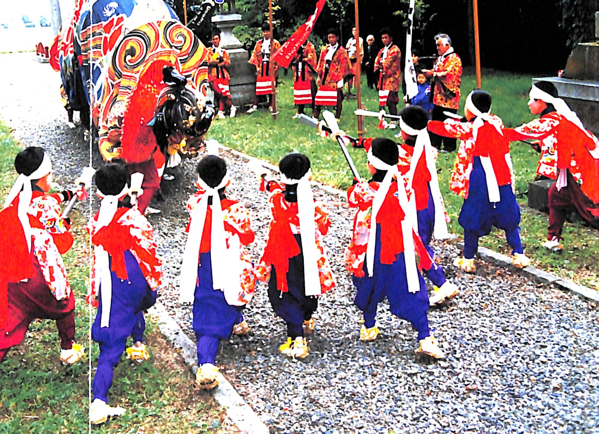 神社内の石を敷き詰めた通路で、手前にいる白い鉢巻で赤い衣装を着た子供たちが手に棒を持ち、奥に獅子舞をけん制しているような様子が写る、砺波獅子舞の写真