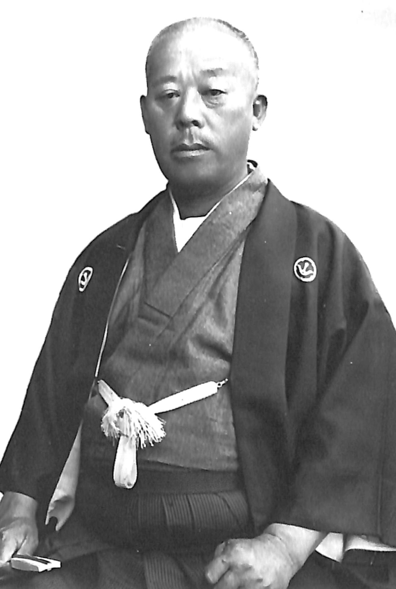 紋付き袴で右手に閉じた扇子を持ち座っている、山田勢太郎のモノクロ写真