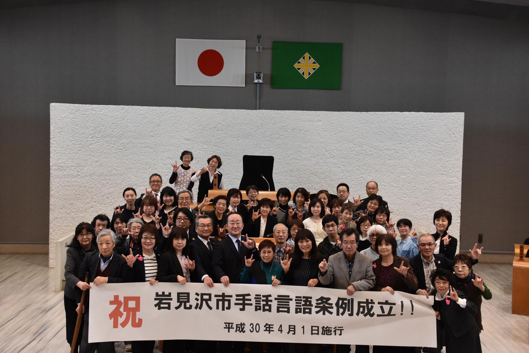 大勢の人たちが演台の前で横断幕に手をかけ集合している様子の写真