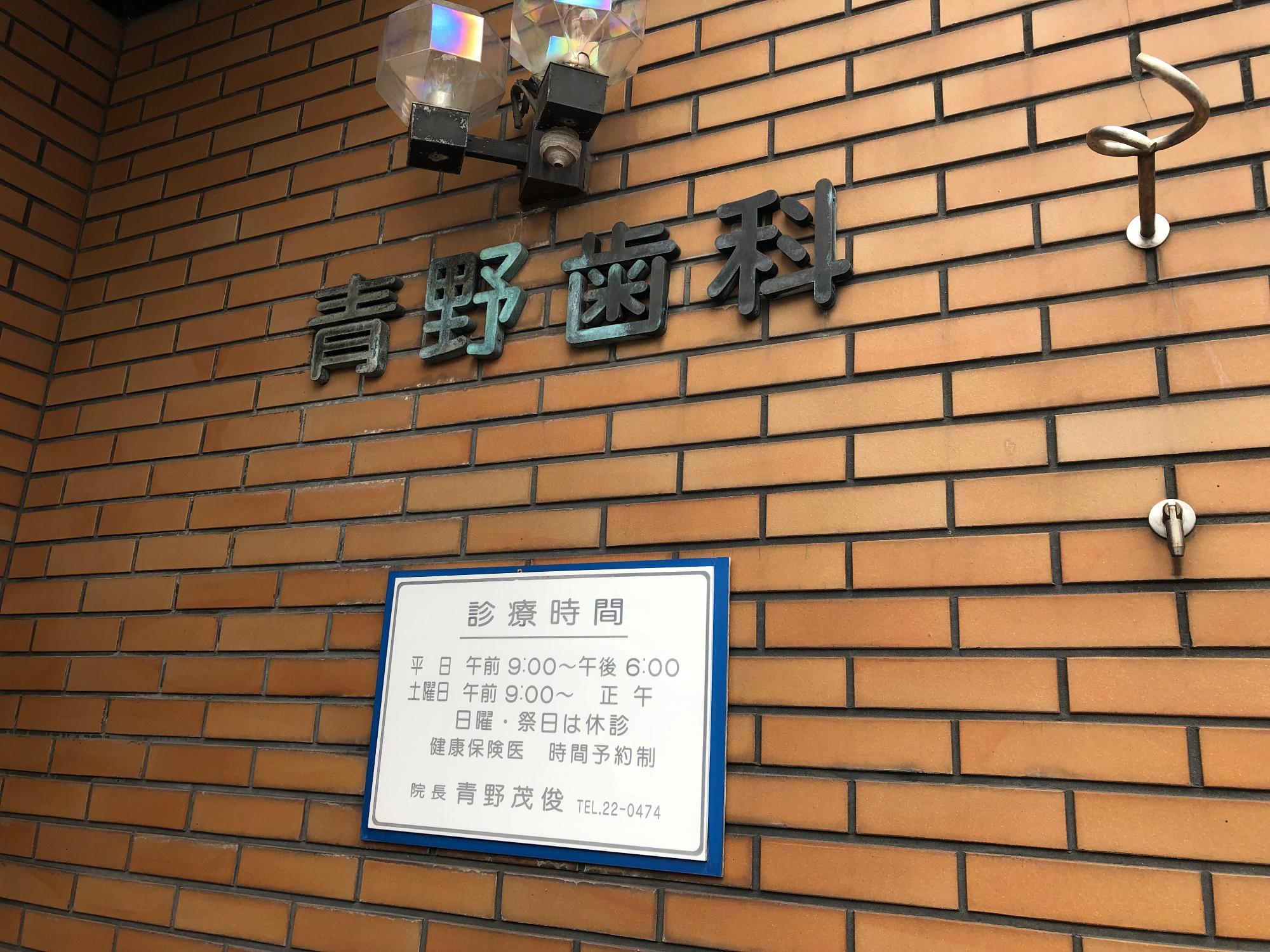 レンガ風の壁に青野歯科の文字看板と診療時間の書かれた看板が並んでいる写真