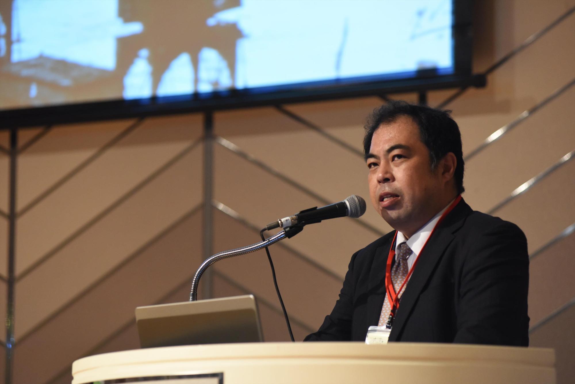 講壇に設置されたマイクを使い挨拶をするスーツ姿の西川賢司さんの写真