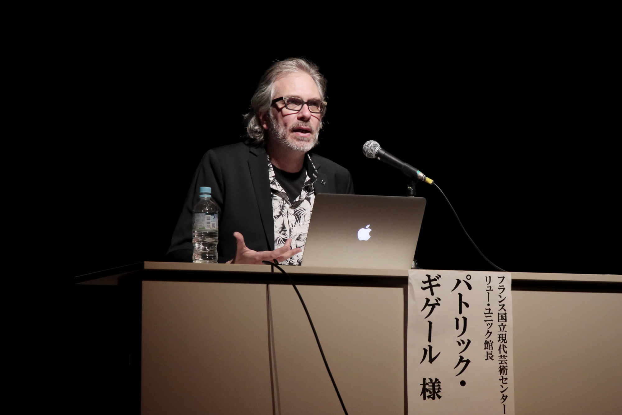 スポットライトが当たった舞台上のテーブルに向かい、パソコンとマイクの前で話をする外国人男性の写真