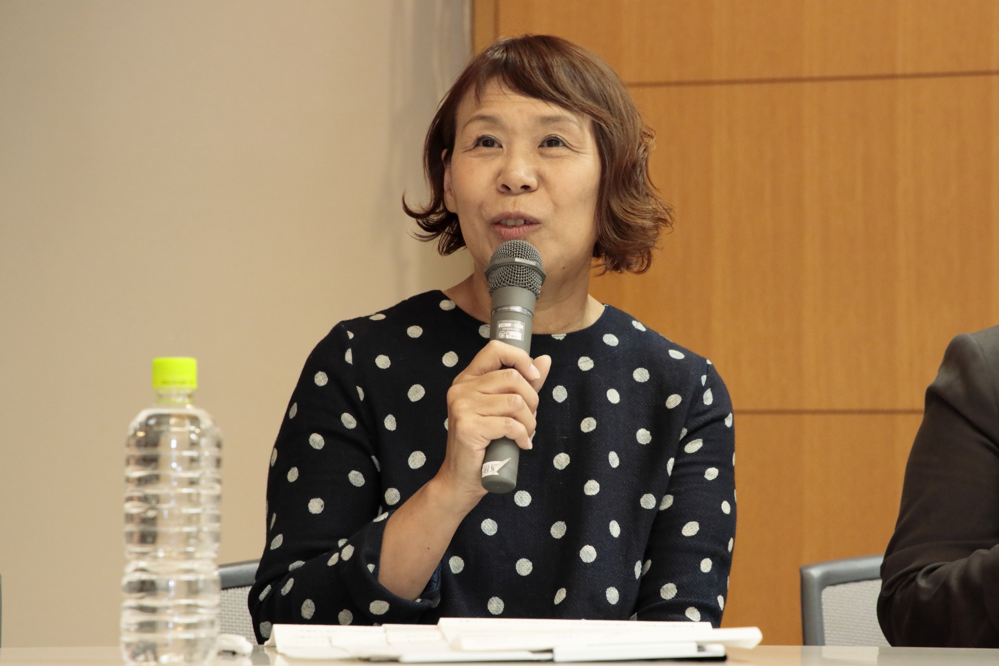 室内の机に向かいパソコンの前でマイクを持ち話をする、水玉模様の洋服を着た女性の写真