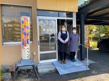 入口の前にこちらを向いたお店の人が2人並んで立っているサンライズ美容室の入口の写真