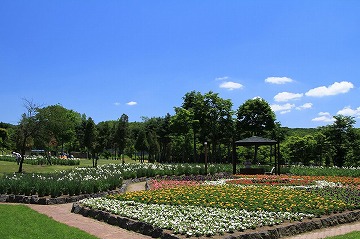 背景に青空、手前にさまざまな花や咲いている花壇や四阿が写っている写真