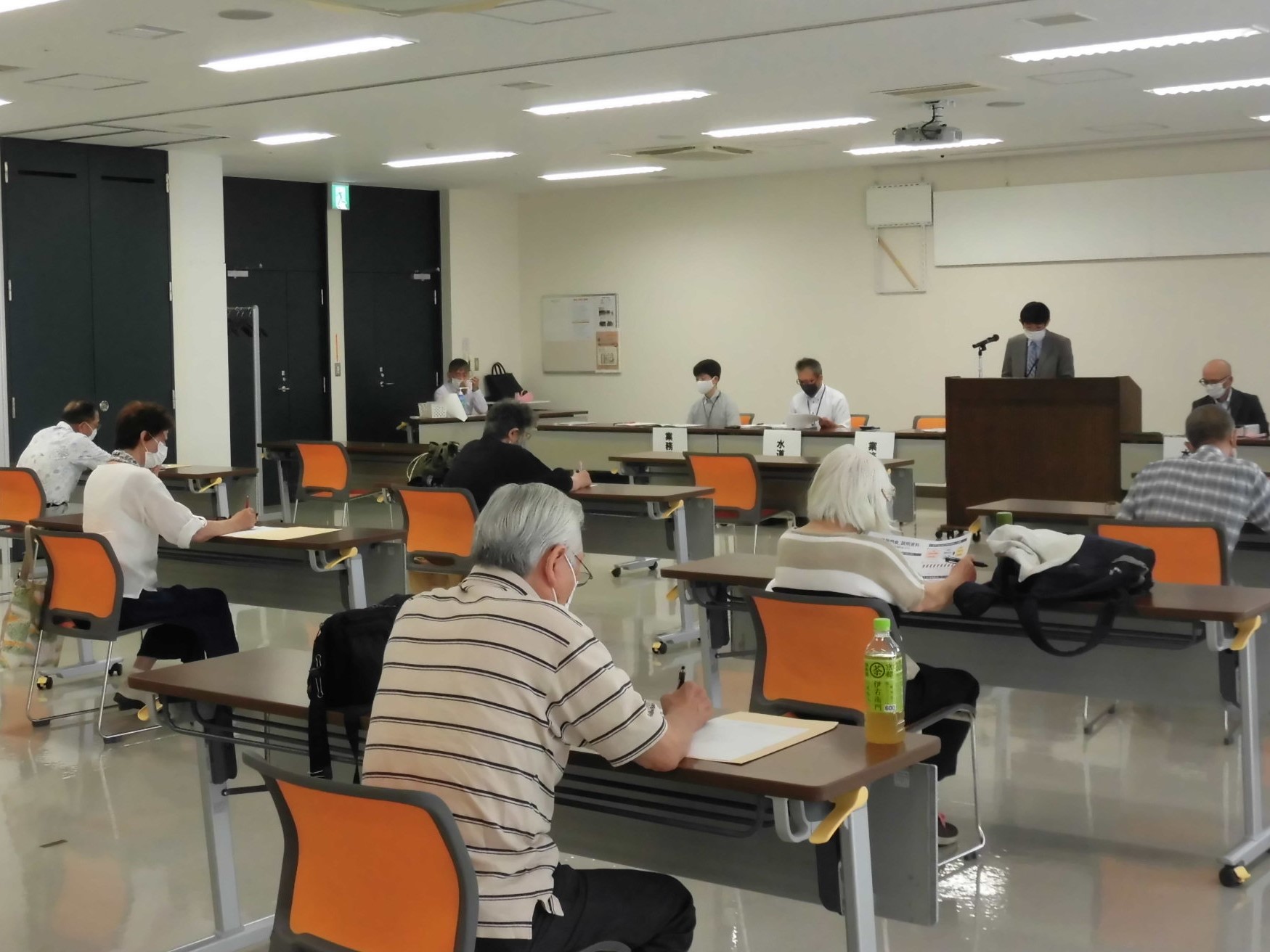 奥にワイシャツを来た男性3人とスーツを着た男性2人がいて、右から2番目の男性はマイクの前に立っていて、手前に6人が椅子に座って話を聞いている写真