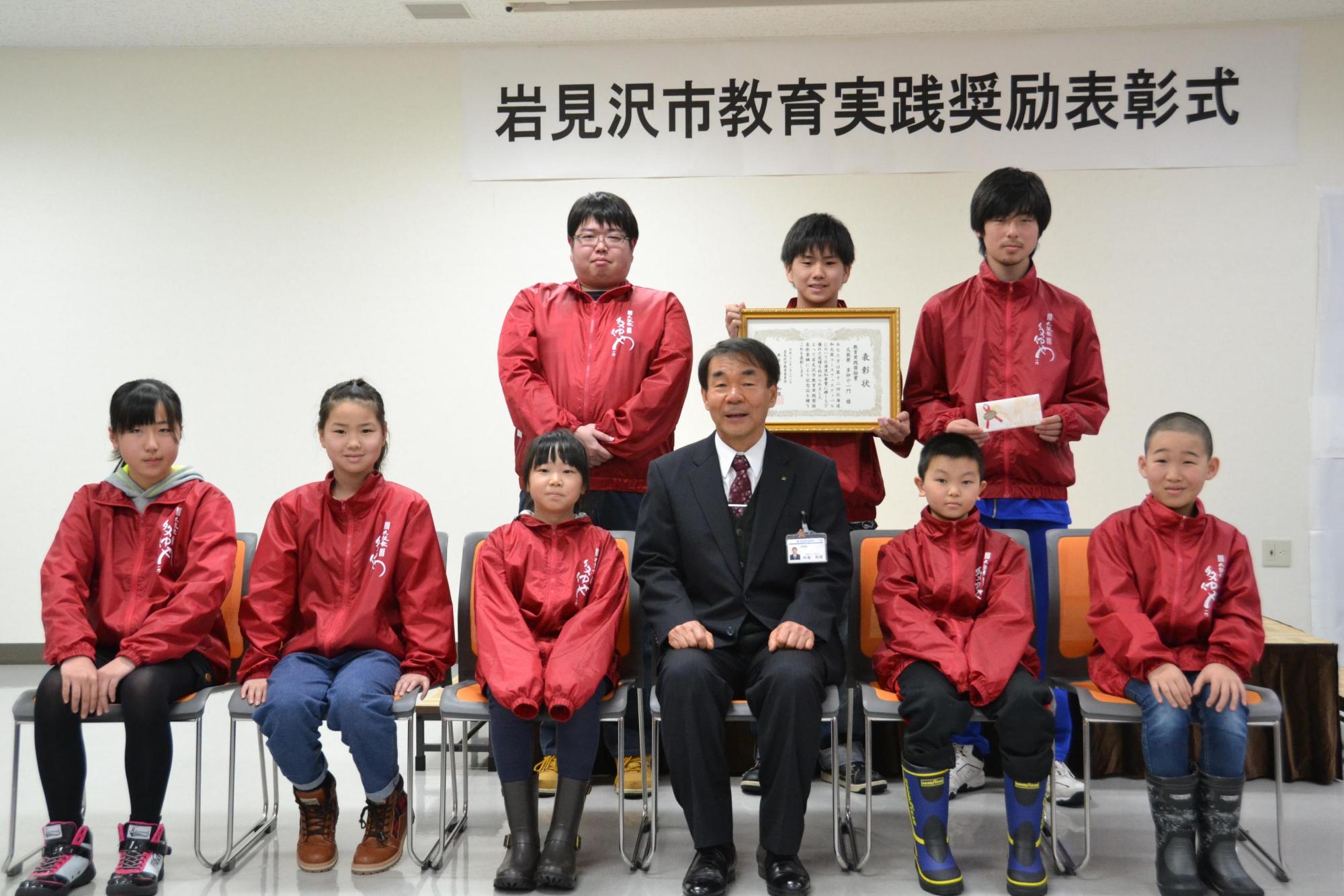 赤のジャンパーを着た受賞者の皆さんが教育長を囲んで記念撮影している写真