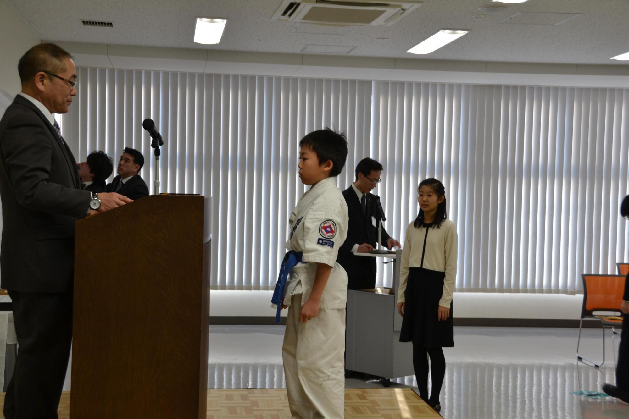 壇上にはスーツを着て眼鏡をした男性と道着の男子児童が立っている室内で行われた表彰式の写真