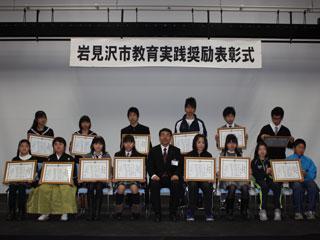 表彰式で座る小学生と中学生の男女と大人の男性の写真