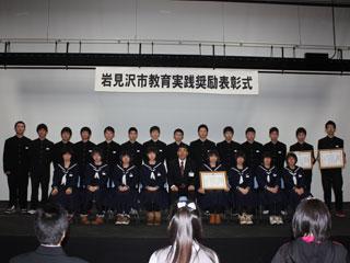 表彰式で立った団体の中学生の男子と椅子に座る女子と一人の男性の様子の写真