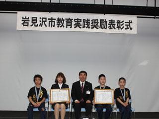 表彰式で座る団体の5人の男女と中央の男性の様子の写真