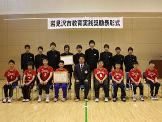 団体受賞者の小中学校の表彰式に立つ数人の男女と椅子に座る数人の男女の様子の写真