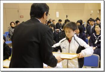 賞状を手渡す職員とスポーツウエアを着て受けとる小学生の写真