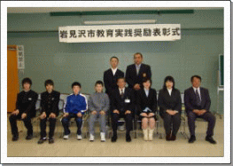 団体受賞者の表彰式で立つ二人の男性と座る数人の男女の様子の写真