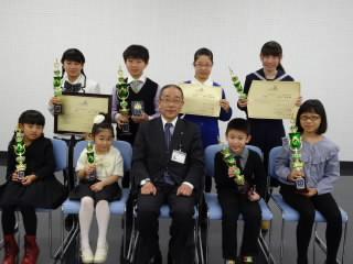 前列に教育長を真ん中にして左右2人の生徒がトロフィーを抱えて座り、後列に賞状とトロフィーを抱えた4人の生徒が並んでいる記念写真