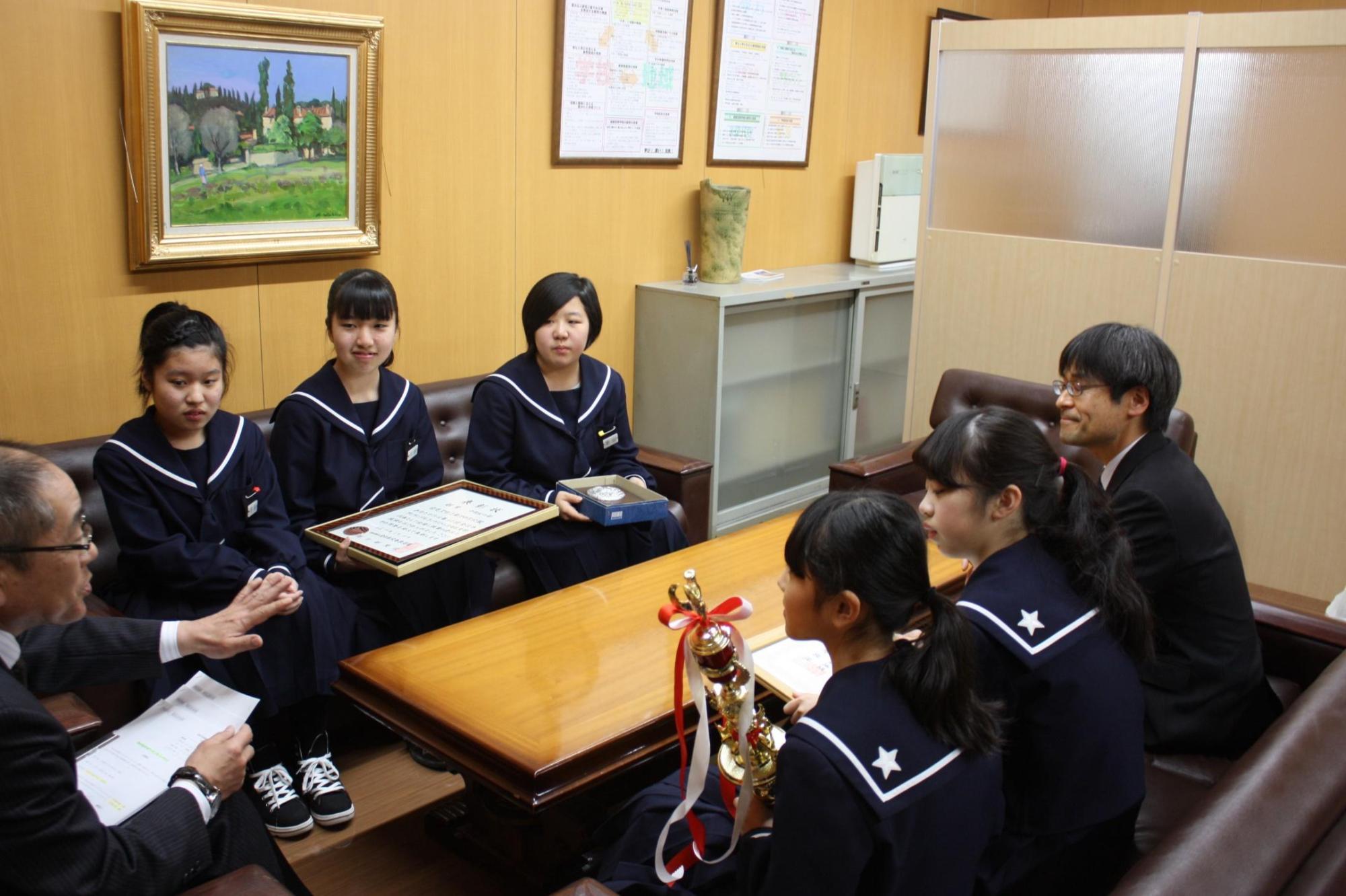 教育長と賞状やトロフィーを抱えた5人の女子生徒と1人の男性がテーブルを囲み対話をする様子