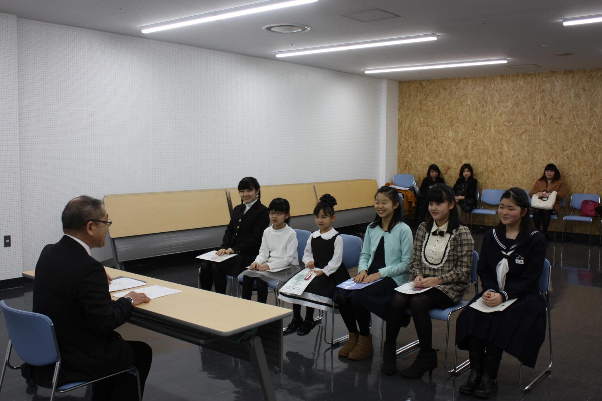 教育長と机を挟んで、6人の入賞者が椅子に座っている様子