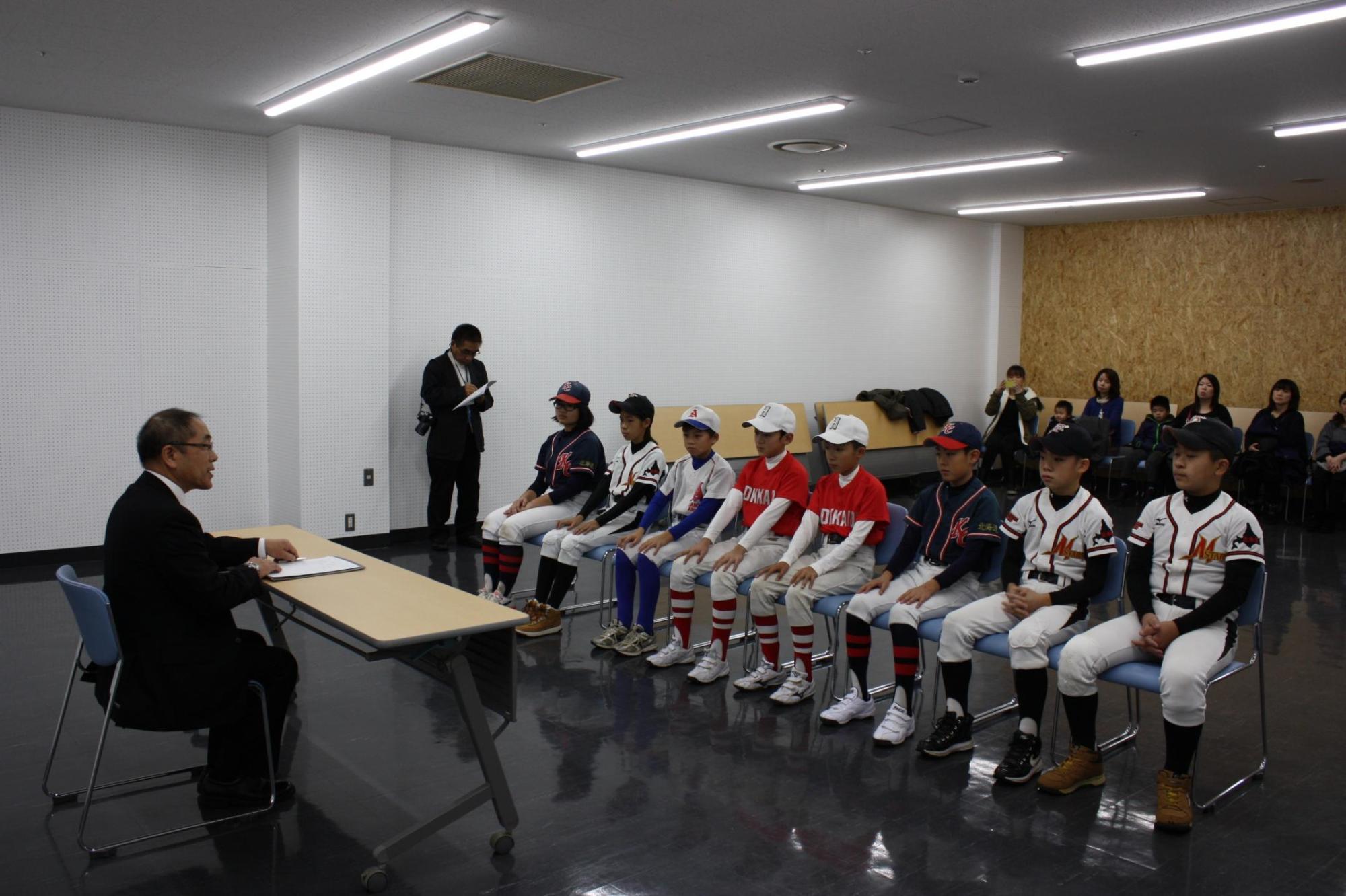 教育長と机を挟んで、8人のユニフォーム姿の選手が椅子に座っている様子