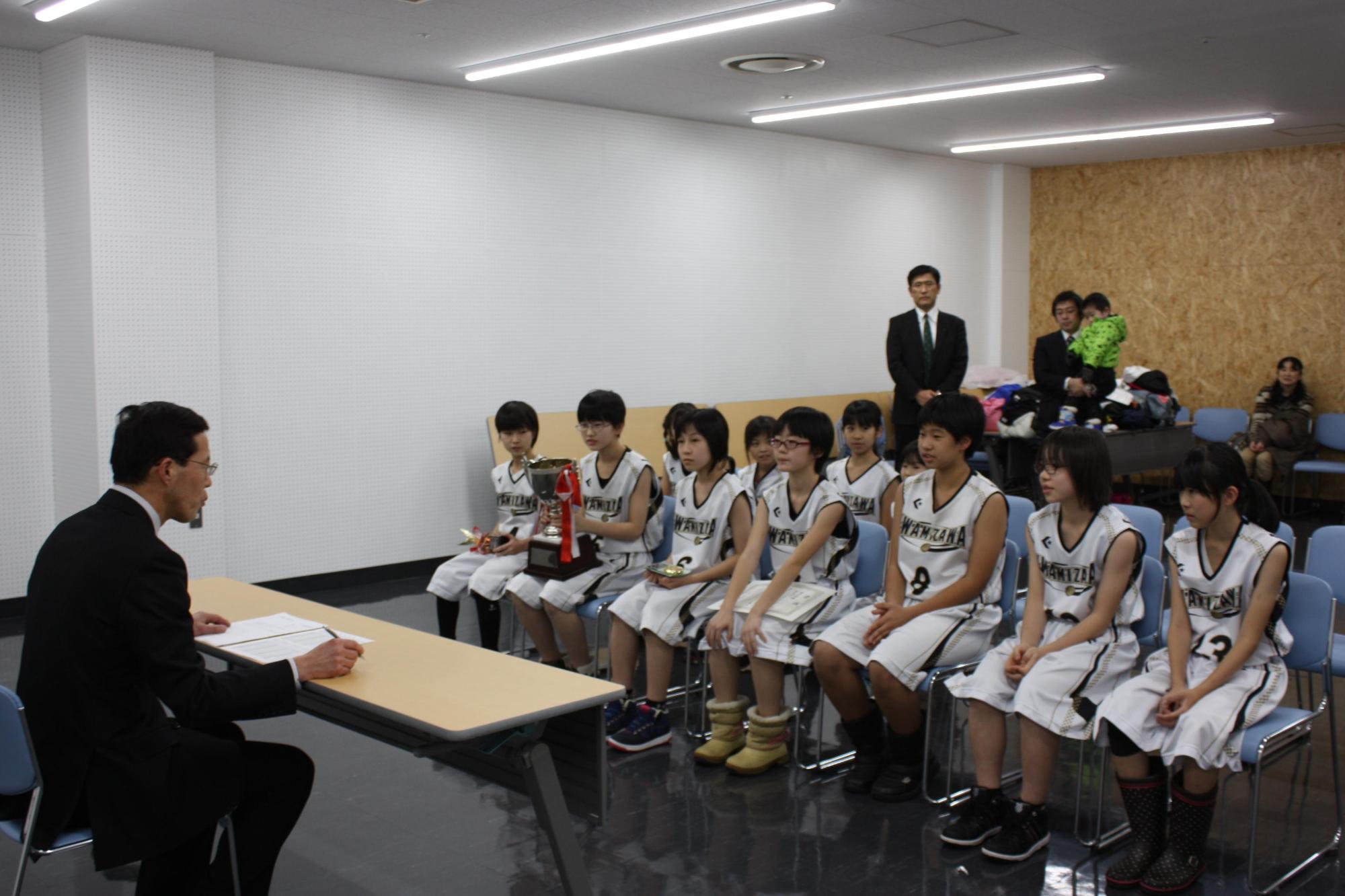 スーツを着た男性と机を挟んで、ユニフォーム姿の11人の選手が椅子に座っている様子
