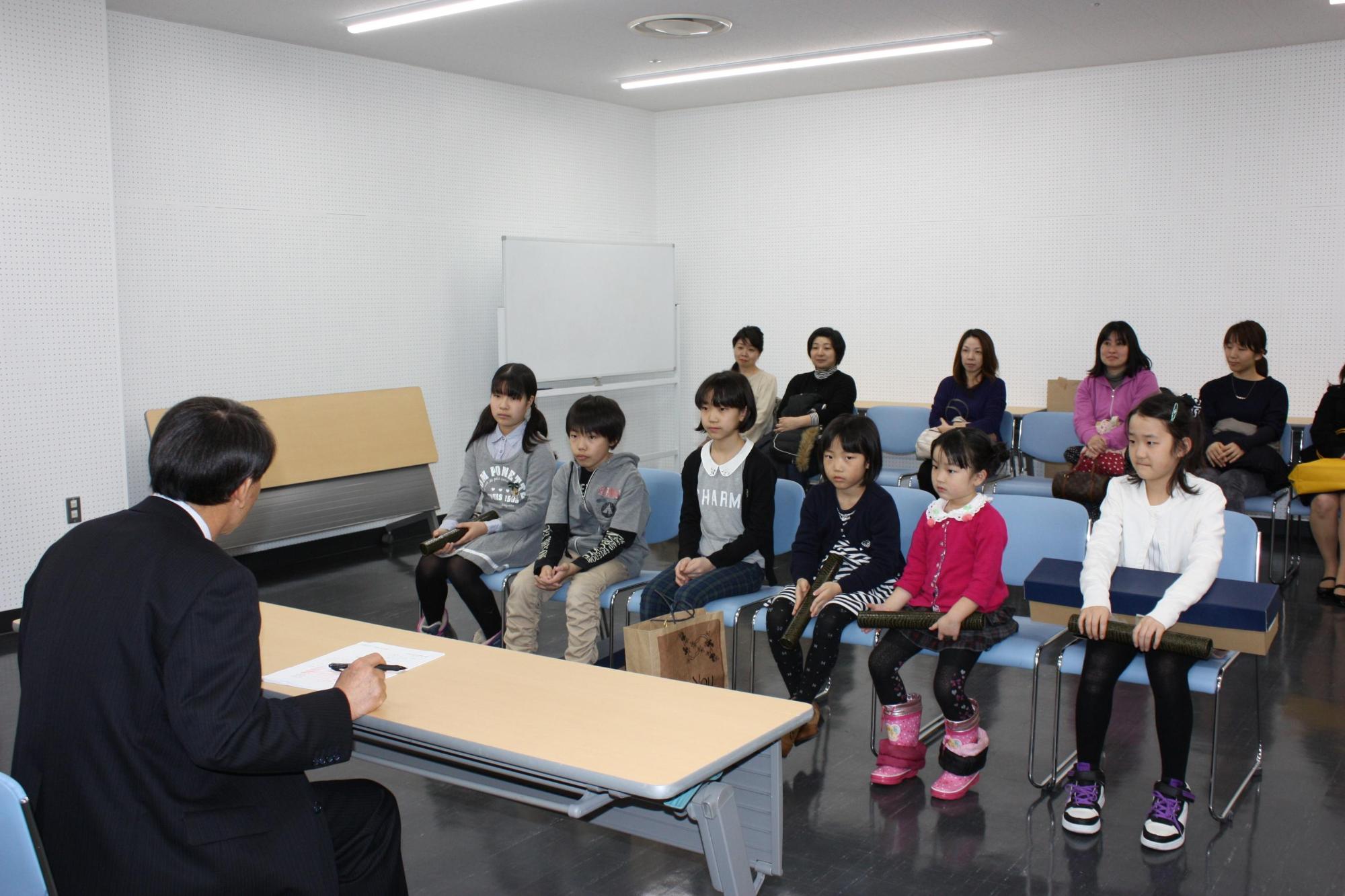 話をする教育長と、その話を聞く6人の子供達と保護者の写真。