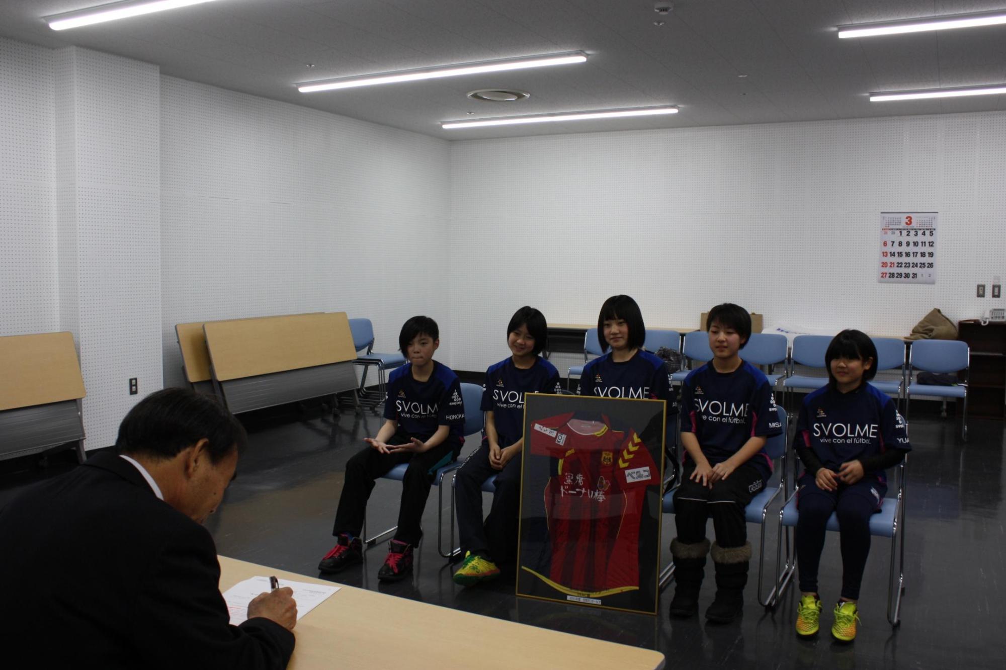書類に記載をする教育長と、その様子を見守る5人の子供達の写真。