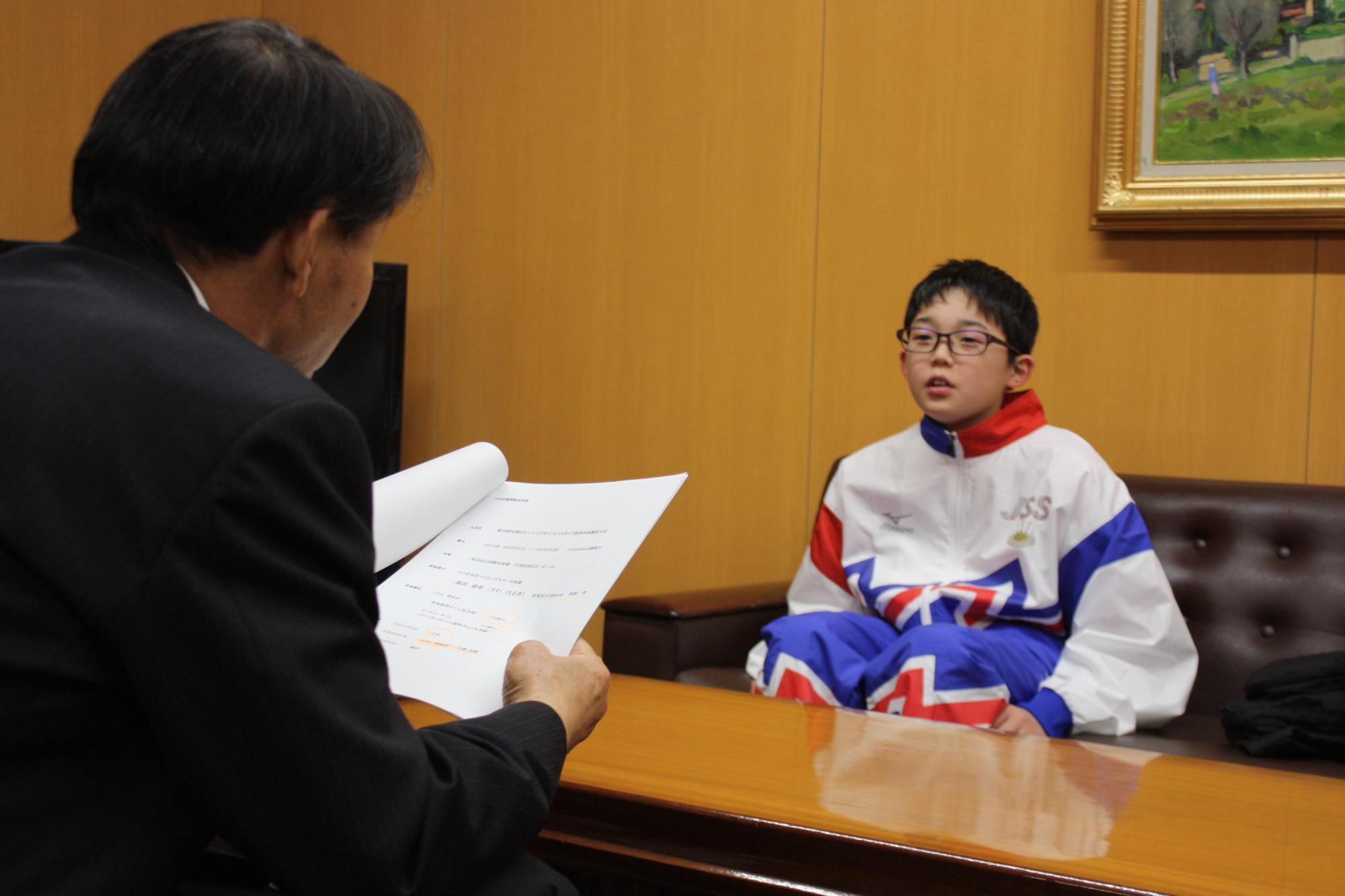 書類を手にした教育長と、ウィンドブレーカーを着た子供が机を挟んで話をしている様子の写真。