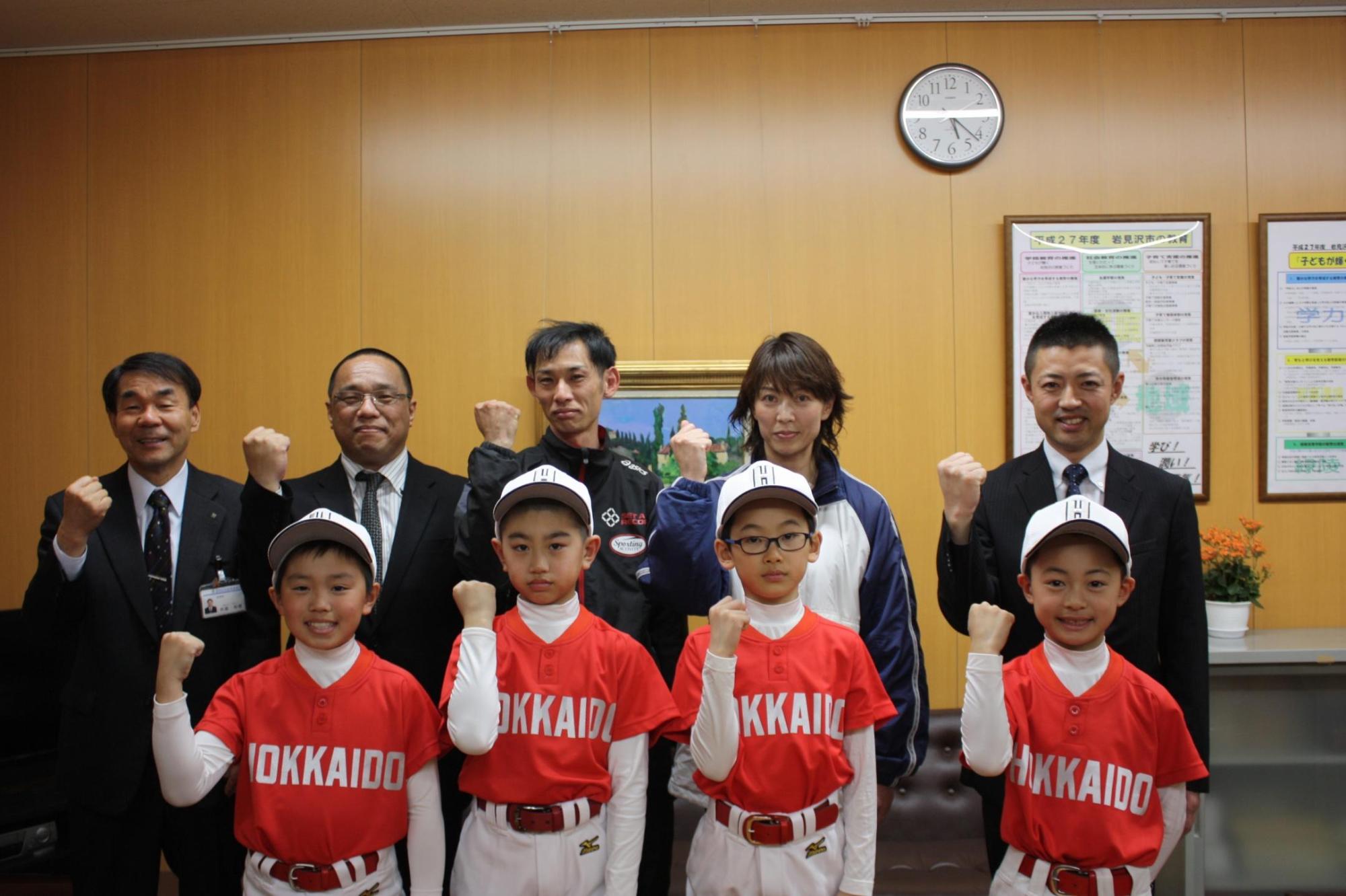 野球のユニフォームを着た4人の子供達と、5人の大人達がガッツポーズをして記念撮影をした写真。