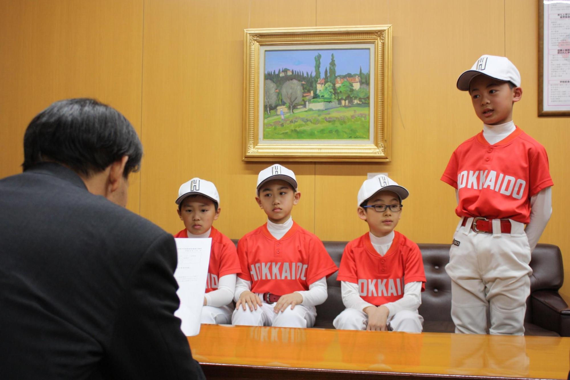 4人のユニフォームを着た子供達と、教育長が机を挟んで話をしている写真。