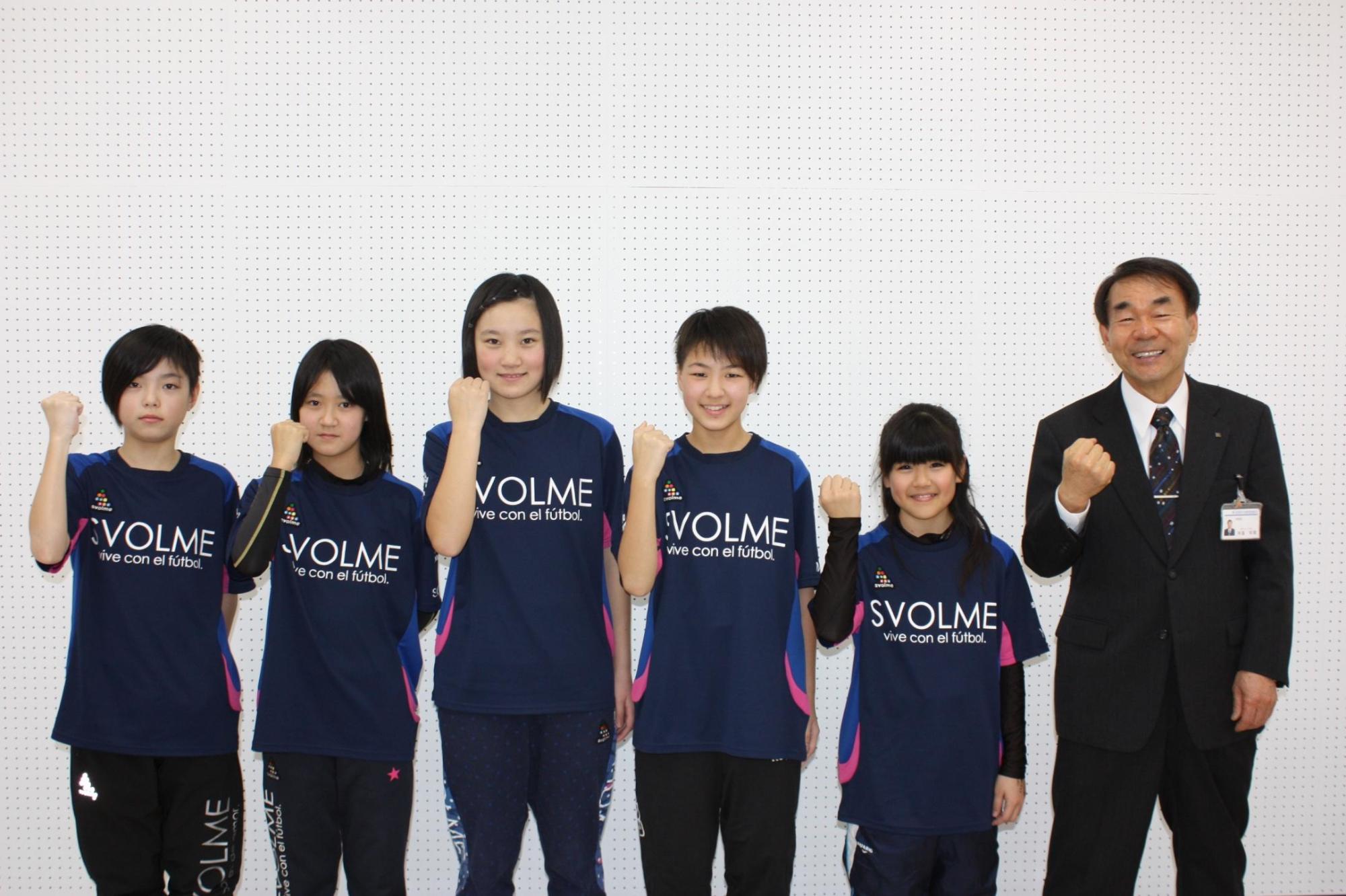 サッカーのユニフォームを着た5人の女の子と教育長がガッツポーズをして記念撮影をした写真。