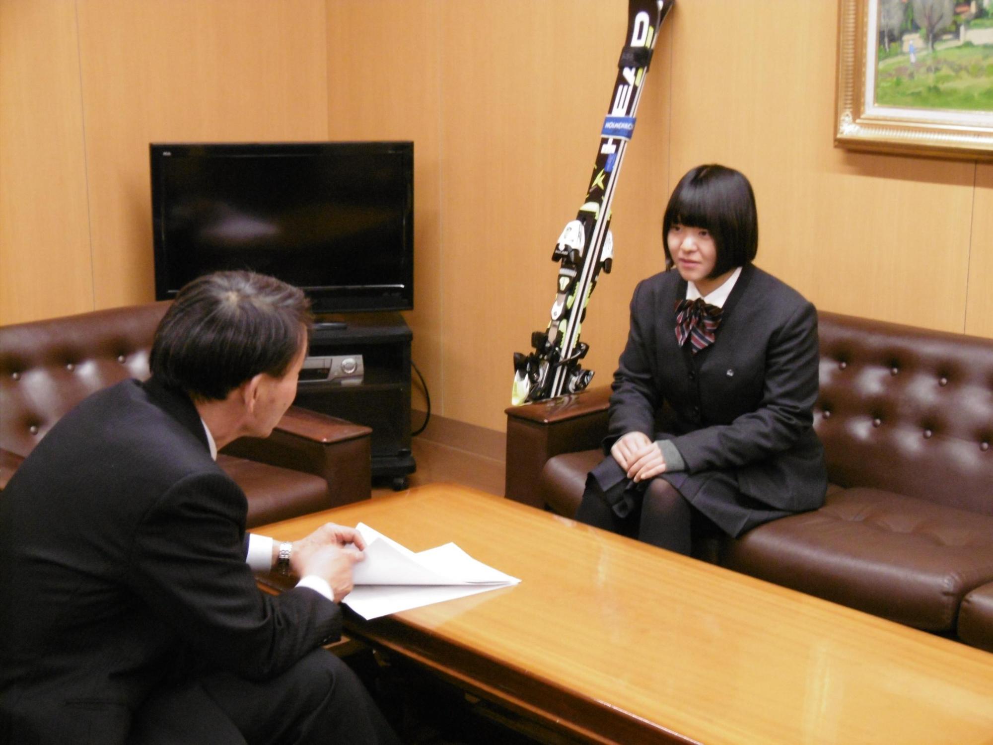 机を挟んで教育長と女子高生が話をしている写真。