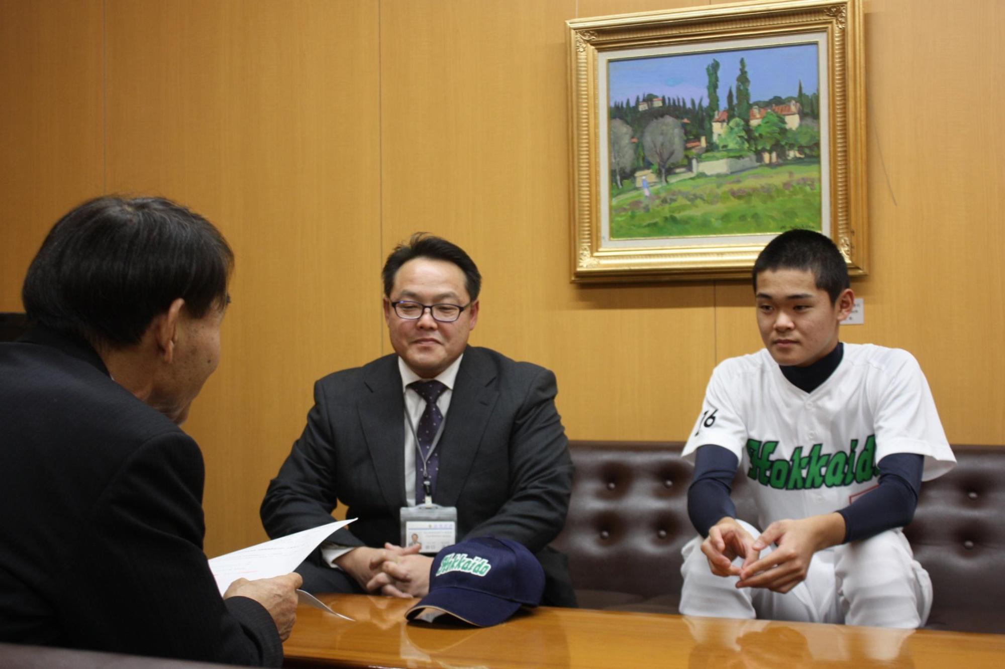 教育長と、野球のユニフォームを着た子供とスーツを着た大人が机を挟んで話をしている写真。