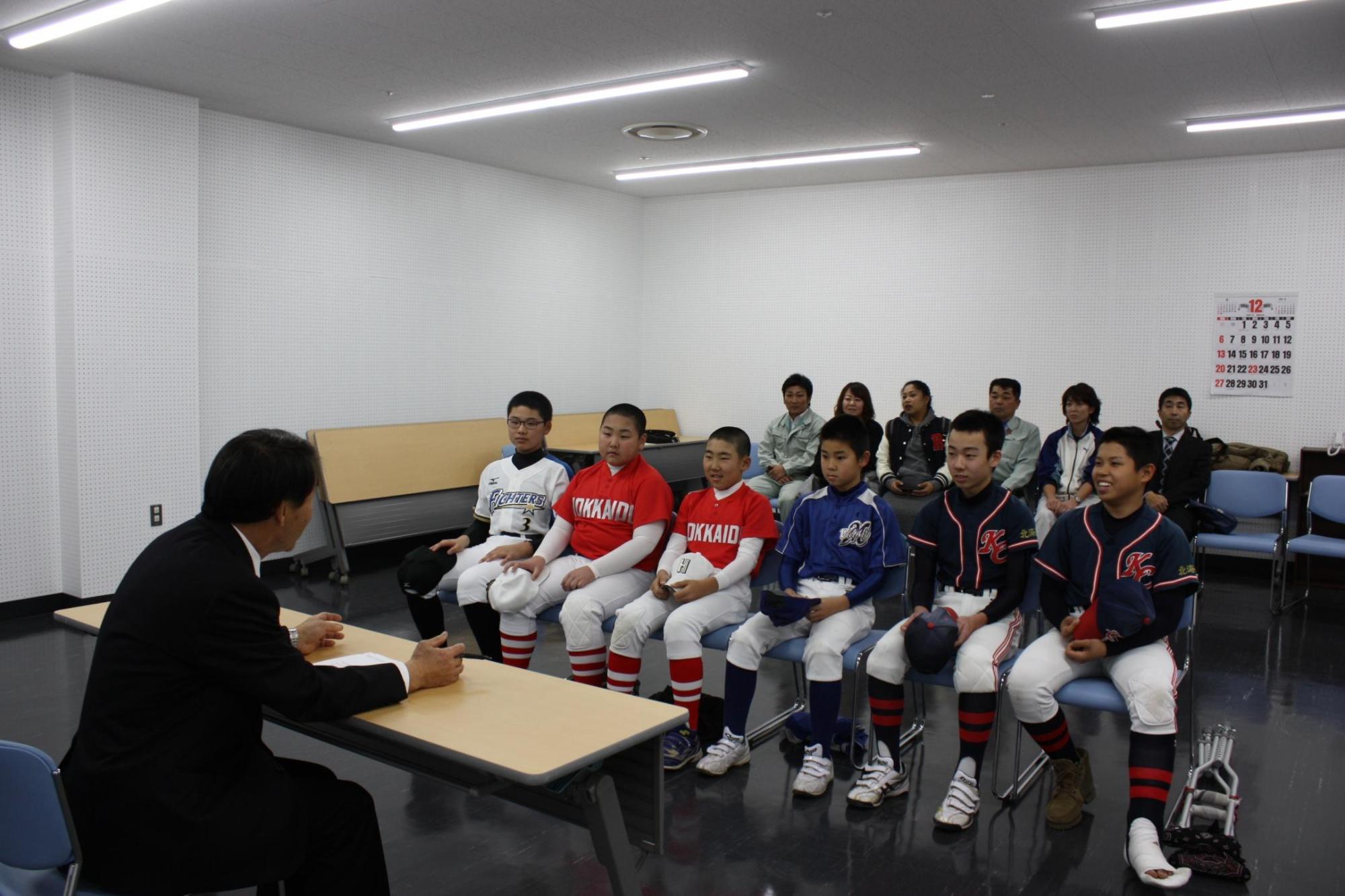 バスケットボールのユニフォームを着た子供数人と保護者が机を挟んで教育長と話をしている写真。