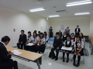 制服を着た学生数人が、机を挟んで教育長の話を聞いている写真。