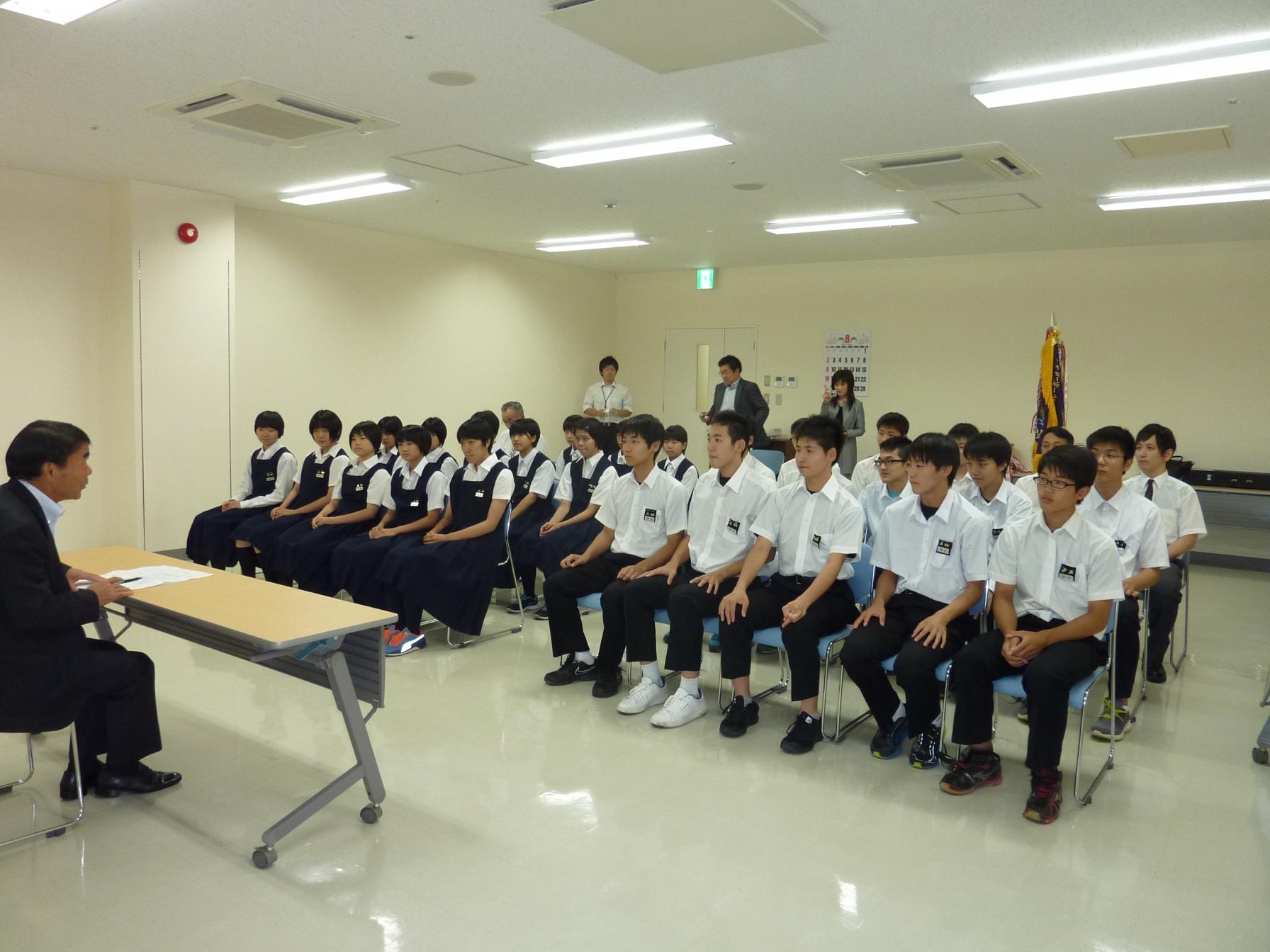 制服を着た学生数人が、机を挟んで教育長の話を聞いている写真。