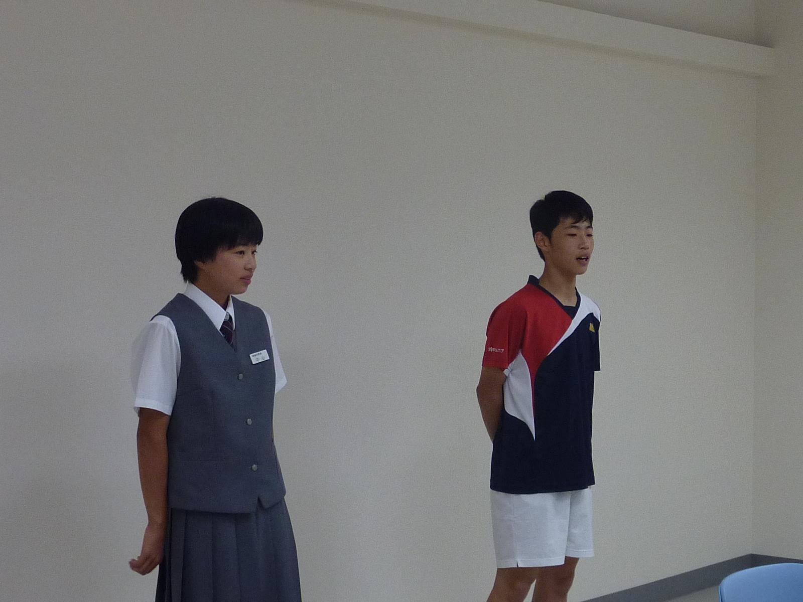 桜本 倖辰さんと制服を着た女の人は白い壁の前に立って発表している写真