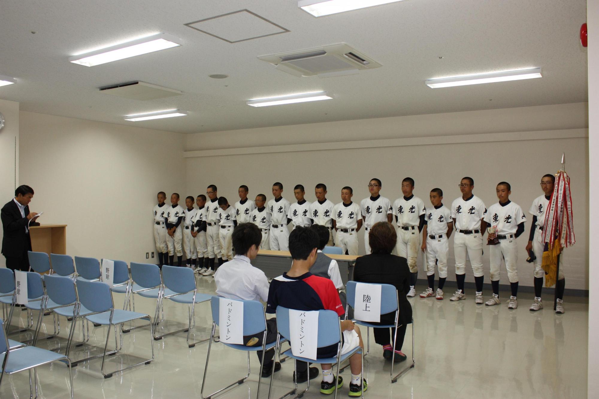 野球のユニフォームを着た子供数人は机を挟んで教育長の話を聞いている写真。