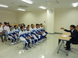 野球のユニフォームを着た学生数人と保護者たちが、机を挟んで教育長の話を聞いている写真。