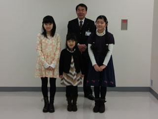 3人の女子生徒が並び、その後ろにスーツを着た男性が立っている写真