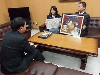 スーツを着た男性と机を挟んで、制服を着た女子生徒が賞状を持ち、スーツを着た男性が絵画を持っている写真