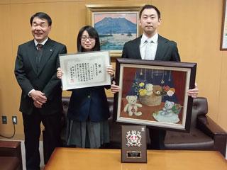 右から、絵画を持ったスーツを着た男性と、賞状を持った制服を着た女子生徒と、手を前で組んでいるスーツを着た男性が並んでいる写真