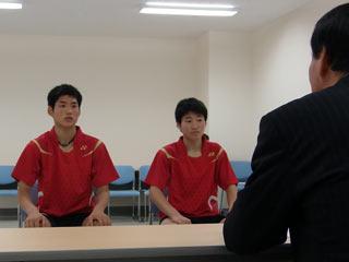 スーツを着た男性と、2人の赤いユニフォームを着た男子生徒が向かい合って座っている写真
