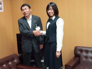 右から、制服を着た女子生徒と、スーツを着た男性が並んで握手をしている写真
