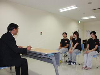 スーツを着た男性と机を挟んで、3人の女子選手と、奥に1人の男性が椅子に座っている写真