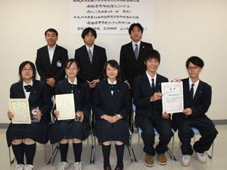 前列、左から制服を着た3人の女子生徒と2人の男子生徒が並んで座っていて、後列はスーツを来た男性3人が並んで立っている写真