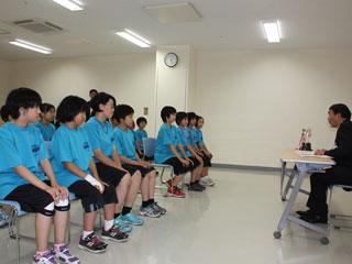 ユニフォームを着た女子生徒達が椅子に座っていて、向かい合わせでスーツを着た男性が椅子に座っている写真