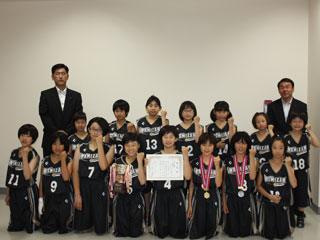 ユニフォームを着た女子生徒達15名が地面に座っていて、奥にスーツを着た男性2名が立っている写真