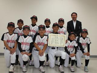 ユニフォームを着た前列6人、後列5人の子供達が並んで立っていて、前列の右から3番目は賞状をこちらに向けており、奥にスーツを着た男性が立っている写真