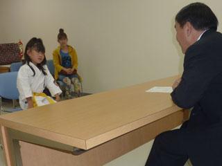 スーツを着た男性と机を挟んで、道着を着た女子生徒と、奥に1人の女性が椅子に座っている写真